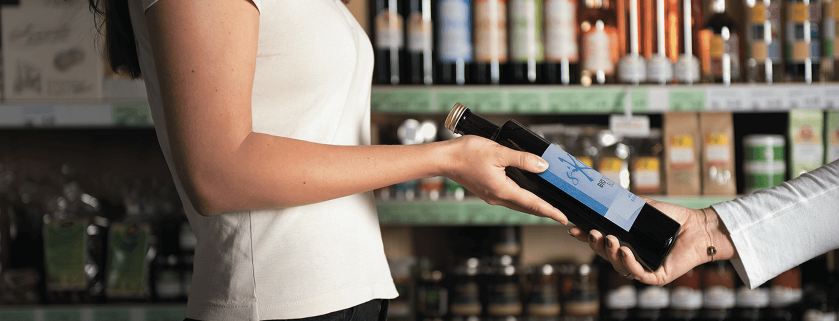 Eine Person überreicht eine Flasche Öl an eine Konsumentin, Regale mit regionalen Produkten